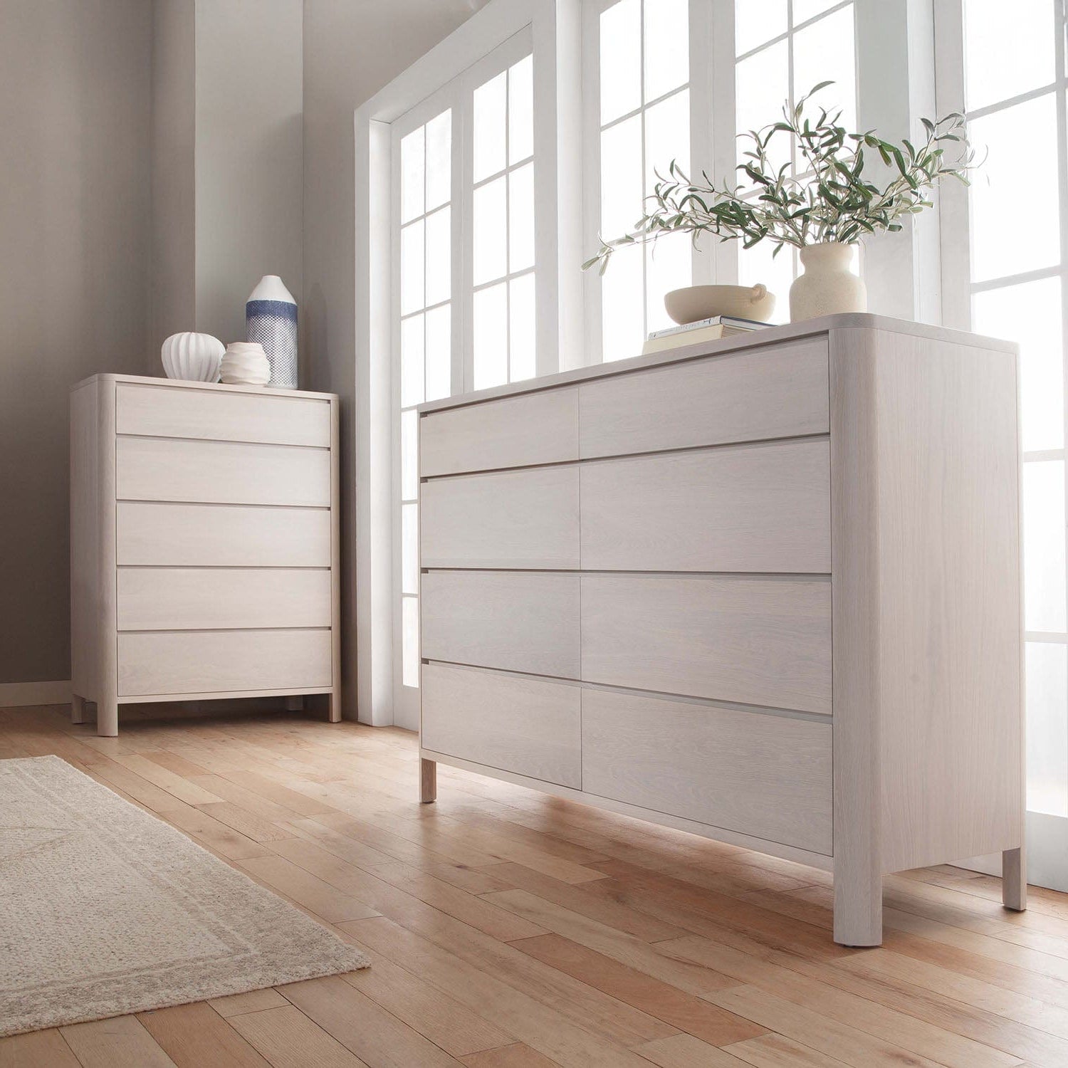 Two light gray dressers in a room with large windows and wooden flooring.