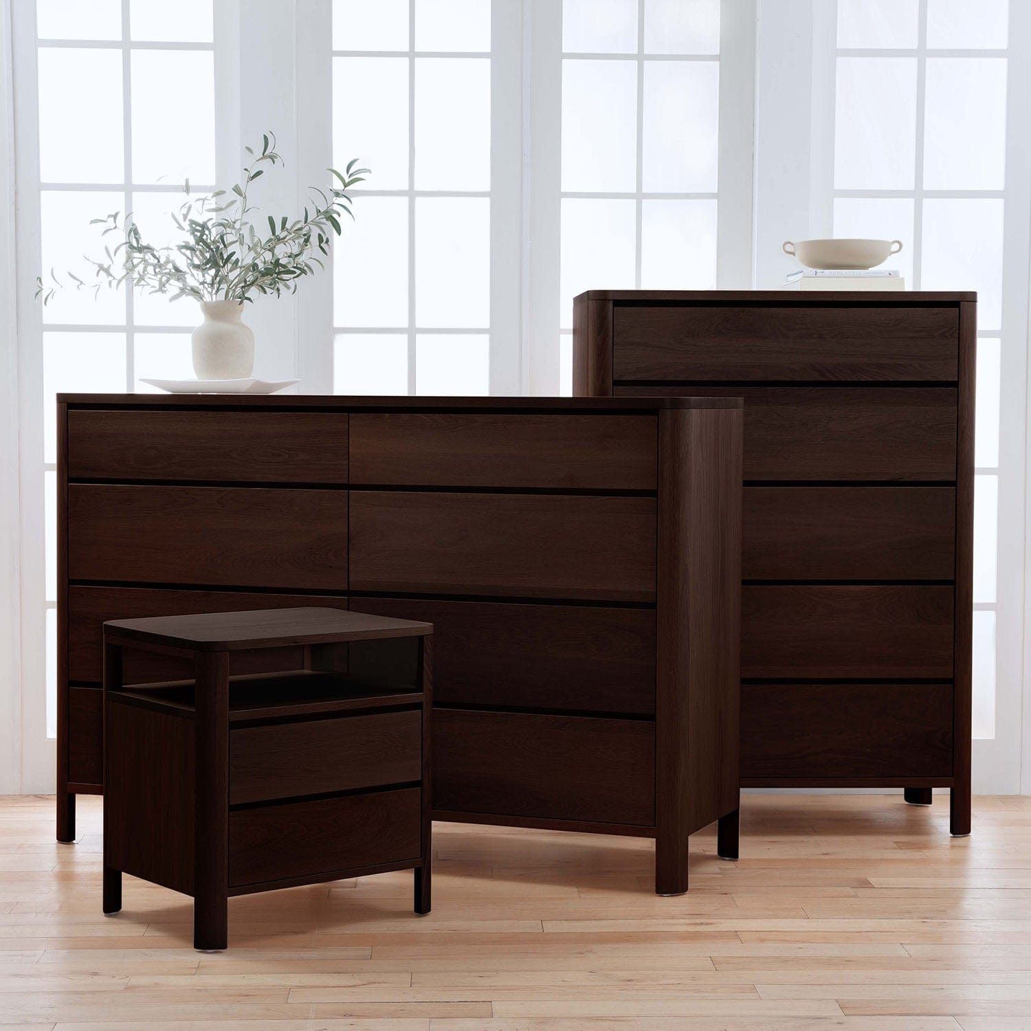 Dark wood bedroom furniture set including a dresser, nightstand, and chair in a bright room.