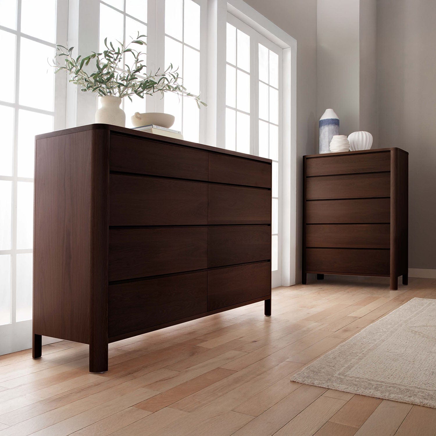 Two dark wooden dressers in a room with large windows and a rug.
