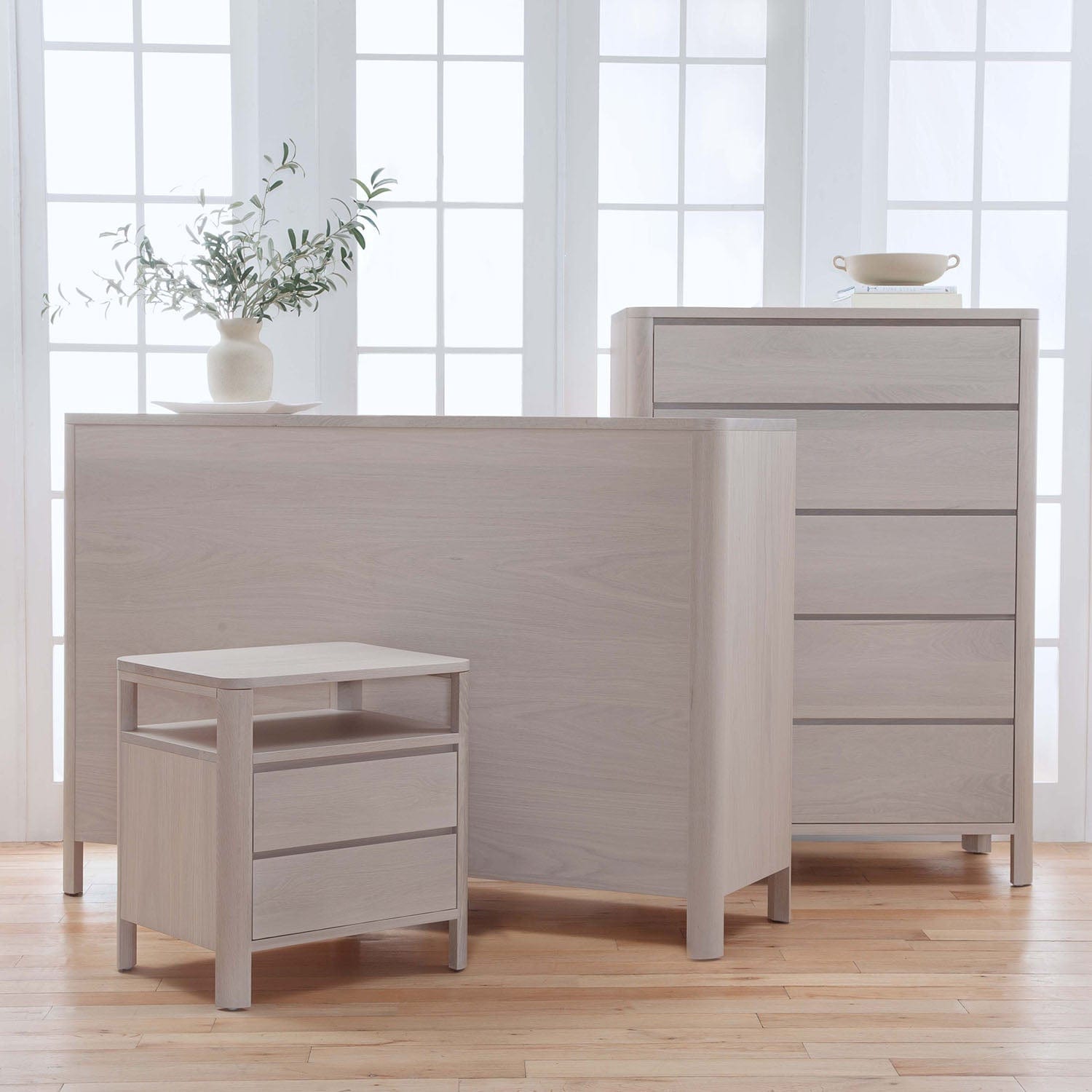Light wood furniture set including a dresser, table, and stool in a bright room with large windows.