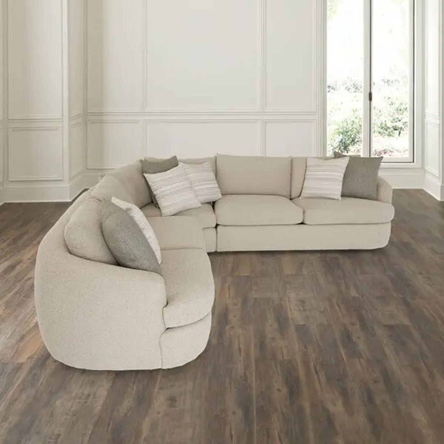 Beige sectional sofa with cushions in a room with wooden flooring and large windows.